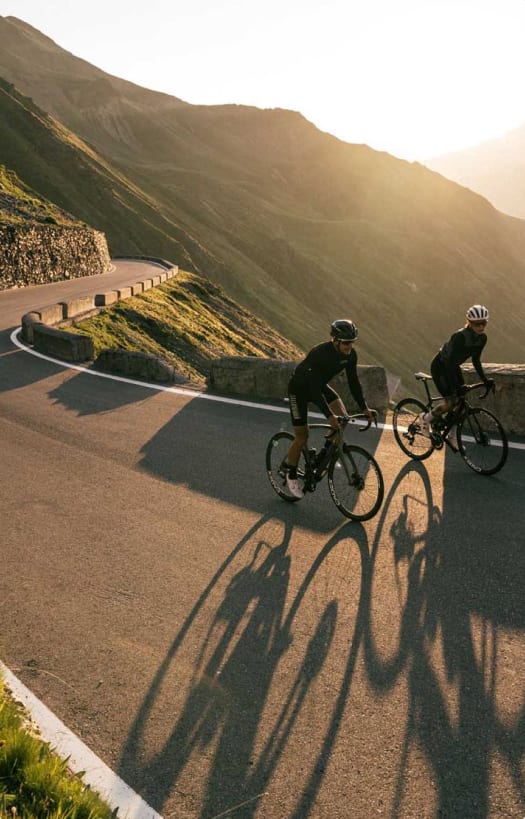 Zwei Rennradfahrer in einer Bergkehre bei Sonnenuntergang